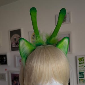 May include: A headband featuring two tall, slender green antennae and two pointed, furry green cat ears. The ears have a gradient effect, transitioning from a lighter lime green at the base to a darker, vibrant green at the tips, with tufts of green fur.