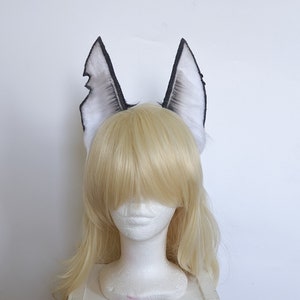 May include: A pair of white and black faux fur wolf ears. The ears are attached to a headband and are worn on a mannequin with blonde hair.