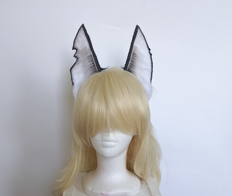May include: A pair of white and black faux fur wolf ears. The ears are attached to a headband and are worn on a mannequin with blonde hair.