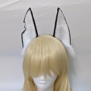 May include: A pair of white faux fur ears with black trim. The ears are attached to a headband and are worn on a mannequin with blonde hair.