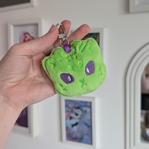 May include: A plush lime green keyring shaped like a cat head with embroidered details. The cat features large purple eyes, a small stitched nose, and whiskers. A purple ribbon loop connects the plush to a silver-toned metal lobster clasp.
