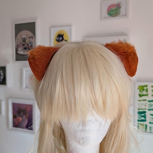 Op de afbeelding: Een blonde pruik met oranje, pluizige oren bevestigd aan een witte mannequinhoofd. De pruik heeft lange, gelaagde pony's en een lichte golf. De oren hebben een warme, roodbruine kleur.
