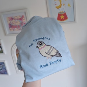 May include: A pale blue hoodie featuring an embroidered pigeon. The pigeon is depicted in shades of grey, white, and pink, with the words "No Thoughts" and "Head Empty" embroidered above and below. The hoodie is folded.