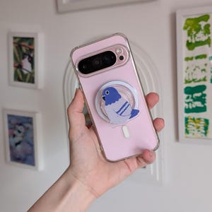 Peut inclure: Une coque de smartphone rose clair avec une poignée circulaire rétractable fixée au dos. La poignée présente une illustration graphique d'un pigeon bleu et blanc. La coque est transparente sur les bords et possède une découpe pour l'appareil photo.