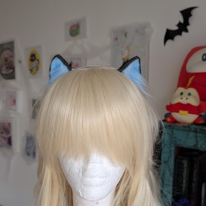 Op de afbeelding: Een blonde pruik met pony op een wit etalagehoofd, voorzien van een paar kattenoren. De kattenoren zijn bedekt met zacht, lichtblauw imitatiebont met een zwarte rand langs de buitenkant, geplaatst bovenop het blonde haar.