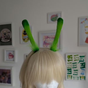 May include: A headband featuring two upright, green, fuzzy, cylindrical antennae. The antennae are covered in a soft, bright green material with darker green tips, attached to a thin, flexible band worn on a blonde wig with bangs.