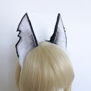 May include: A pair of black and white faux fur ears with a pointed tip. The ears are attached to a headband and are worn on a blonde wig.