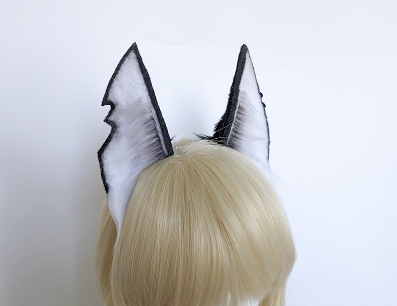 May include: A pair of black and white faux fur ears with a pointed tip. The ears are attached to a headband and are worn on a blonde wig.