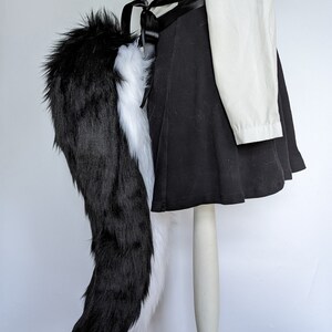 May include: A black and white faux fur tail with a black satin ribbon tied around the base. The tail is attached to a mannequin wearing a black skirt and a white shirt.