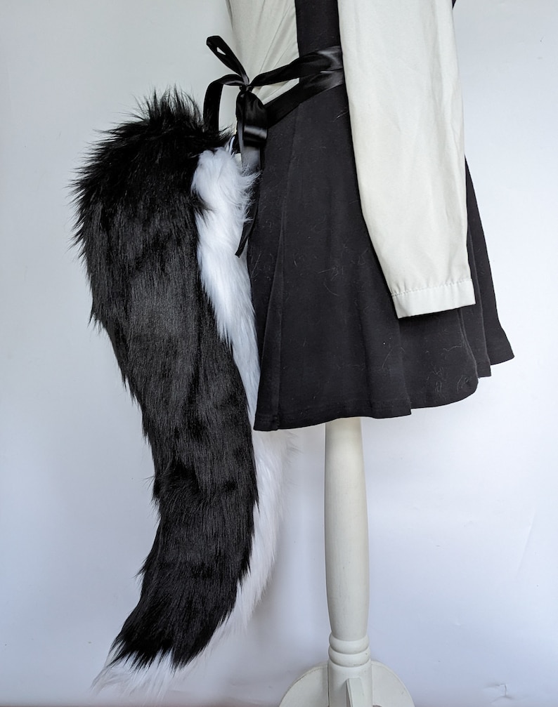 May include: A black and white faux fur tail with a black satin ribbon tied around the base. The tail is attached to a mannequin wearing a black skirt and a white shirt.