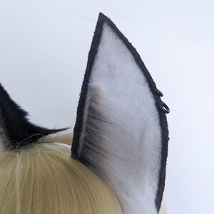 May include: A pair of black and white faux fur cat ears with black elastic bands. The ears are attached to a headband.