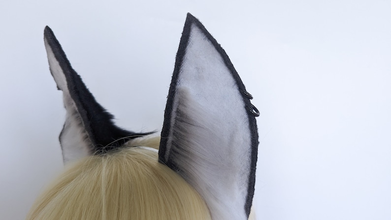 May include: A pair of black and white faux fur cat ears with black elastic bands. The ears are attached to a headband.