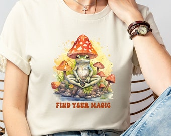 Op de afbeelding: Een wit T-shirt met een aquarel-illustratie van een kikker die onder een paddenstoel zit. De tekst "Find Your Magic" staat onder de kikker.