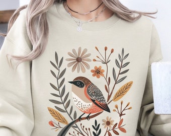 May include: A beige sweatshirt with a colorful bird and floral design. The bird is brown, orange, and gray with a long tail. The flowers are pink, yellow, and orange. The text "SAND" is printed on the bottom right corner of the image.