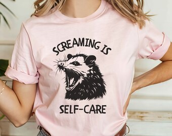 May include: A soft pink t-shirt with a black and white illustration of a possum with its mouth open, and the text "Screaming is Self-Care".