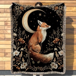May include: A tapestry throw blanket with a black background featuring a stylised orange fox sitting under a crescent moon with gold accents. The fox is surrounded by gold and white floral designs.