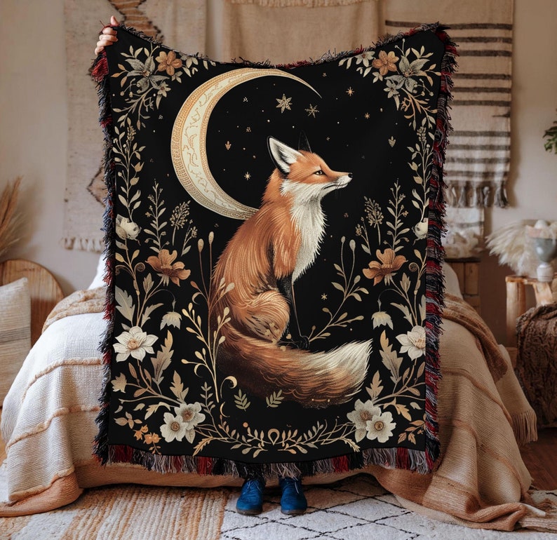 May include: A black throw blanket featuring a gold crescent moon and a fox sitting in front of it. The fox is orange and white with a bushy tail. The background is a dark blue with gold stars and floral designs.