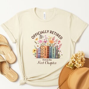May include: A beige t-shirt with the text "Officially Retired & on to the Next Chapter" printed on it. The text is surrounded by a stack of books with floral illustrations.