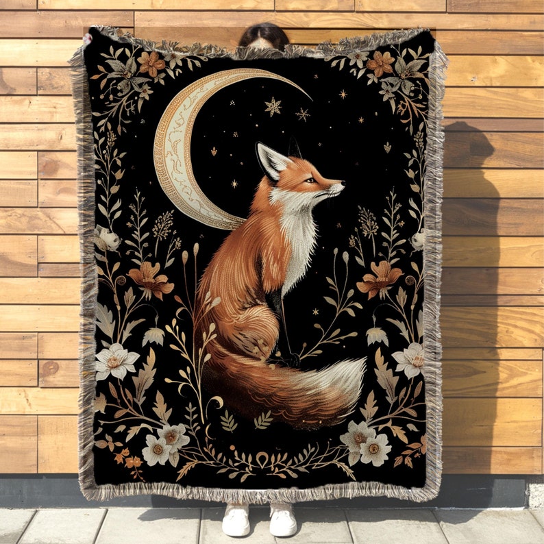 May include: A tapestry throw blanket with a black background featuring a stylised orange fox sitting under a crescent moon with gold accents. The fox is surrounded by gold and white floral designs.
