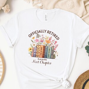 May include: White t-shirt with the text "Officially Retired & on to the Next Chapter" printed in brown. The text is surrounded by a colorful illustration of flowers and books.