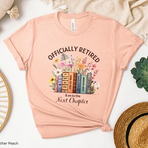 May include: A peach-colored t-shirt with the text "Officially Retired & on to the Next Chapter" printed on it. The text is surrounded by a stack of books with colorful spines and flowers.