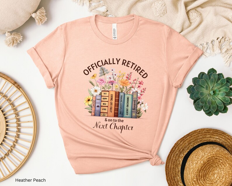 May include: A peach-colored t-shirt with the text "Officially Retired & on to the Next Chapter" printed on it. The text is surrounded by a stack of books with colorful spines and flowers.