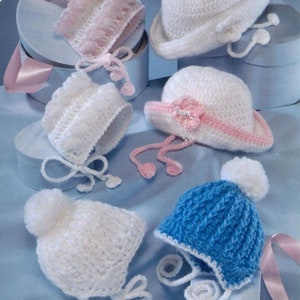 Puede incluir: Cinco gorros de bebé tejidos a crochet en varios colores y estilos. Los gorros se muestran sobre un fondo azul claro con cintas rosas. Los gorros incluyen un gorro blanco con un pompón, un gorro azul con un pompón blanco, un gorro blanco con un borde rosa, un gorro blanco con una flor rosa y un gorro rosa con un borde de encaje blanco.