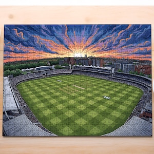 Op de afbeelding: Een rechthoekige legpuzzel met een luchtfoto van een cricketstadion met een groen schaakbordveld, omringd door tribunes en stadsgebouwen onder een levendige zonsonderganghemel met paarse, oranje en blauwe wolken.