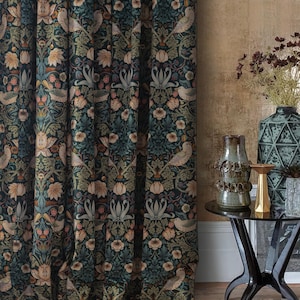 Op de afbeelding: Donkerblauwe gordijn met bloemenpatroon met vogels, bloemen en bladeren. Het gordijn hangt naast een goudkleurige muur. Een kleine zwarte tafel bevat een vaas, een glazen fles en een gouden kandelaar.