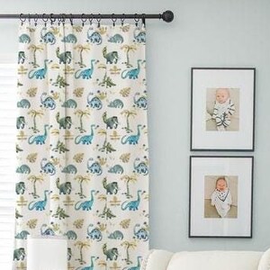 Könnte beinhalten: Ein weißer Kinderzimmer-Schaukelstuhl mit einer grauen Wolldecke, einem weißen Hocker und einem geflochtenen Korb mit einem Hundegesichtsdesign. Der Schaukelstuhl steht vor einem Fenster mit weißen Vorhängen mit einem Dinosauriermuster. Zwei gerahmte Fotos von Babys hängen an der Wand.