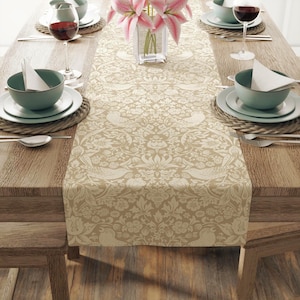 May include: A beige table runner featuring a repeating cream-colored pattern of birds, flowers, and foliage. The runner is centered on a wooden table set with light green bowls, white napkins, wine glasses, and a glass vase with pink lilies.
