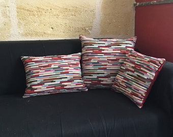 Puede incluir: Tres cojines de sofá rayados de colores sobre un sofá negro. Los cojines tienen un patrón geométrico de rayas horizontales en tonos de rojo, rosa, azul, verde y beige.