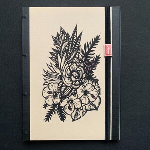 Peut inclure: Un carnet à couverture en papier crème avec une illustration florale noire et blanche. La reliure est noire et porte un tampon rouge avec l'inscription "KA".