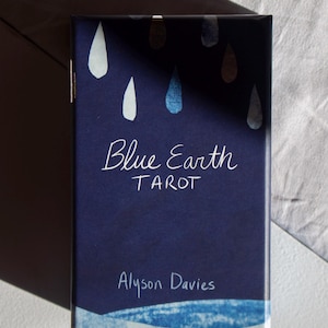 May include: A navy blue tarot card box with the title "Blue Earth Tarot" in white script. The box features watercolor-style teardrop designs in white and blue. The author's name, Alyson Davies, is also printed on the box.