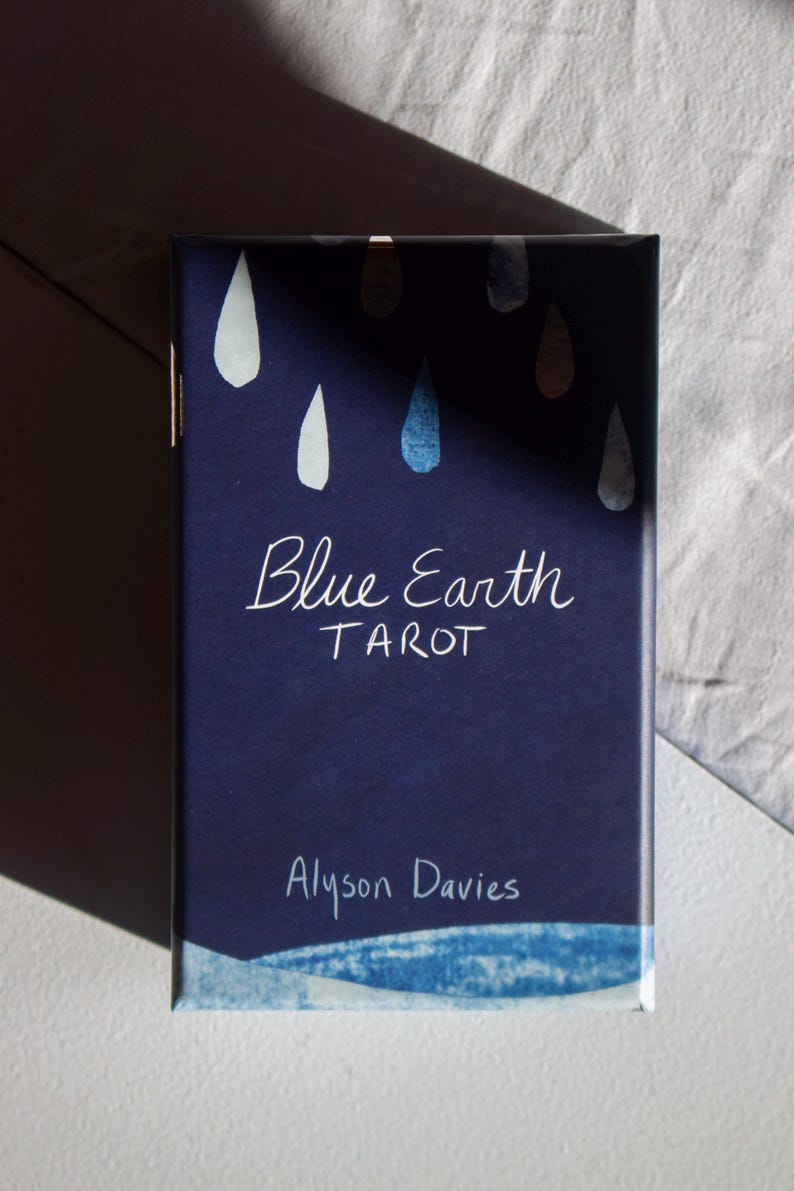 May include: A navy blue tarot card box with the title "Blue Earth Tarot" in white script. The box features watercolor-style teardrop designs in white and blue. The author's name, Alyson Davies, is also printed on the box.