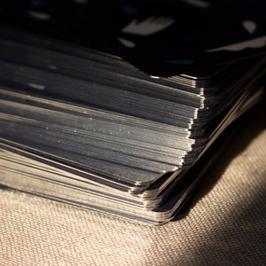 May include: Close-up of a deck of cards with dark blue backs and white and light blue designs. The edges of the cards are silver and reflect light. The deck is resting on a textured, tan surface.