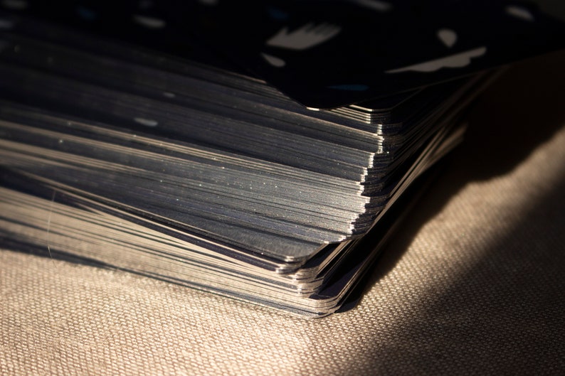 May include: Close-up of a deck of cards with dark blue backs and white and light blue designs. The edges of the cards are silver and reflect light. The deck is resting on a textured, tan surface.