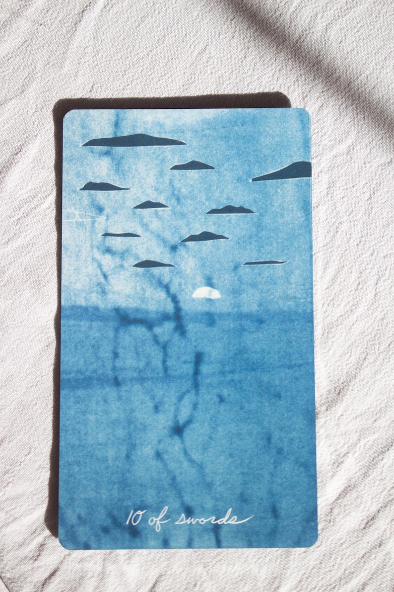 May include: A rectangular tarot card with a blue watercolor design. The card features a sky with cloud shapes and a sun, above a blue sea. The words "10 of swords" are written in cursive at the bottom.