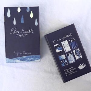 May include: Two navy blue tarot card boxes. One box has the text "Blue Earth Tarot" and "Alyson Davies" with a watercolor design of blue and silver raindrops. The other box has the text "78 cards + guidebook" and a collage of tarot card images.