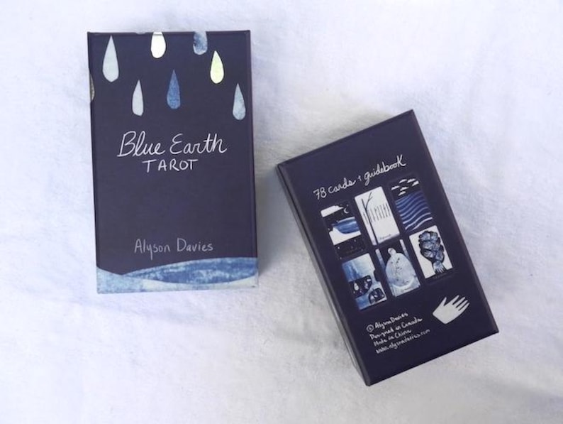 May include: Two navy blue tarot card boxes. One box has the text "Blue Earth Tarot" and "Alyson Davies" with a watercolor design of blue and silver raindrops. The other box has the text "78 cards + guidebook" and a collage of tarot card images.