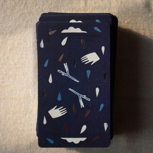 May include: A deck of cards with a navy blue background. The cards feature white hands, clouds, and raindrops in blue and brown. Two stylized branches are also present. The cards are stacked on a neutral-colored surface.