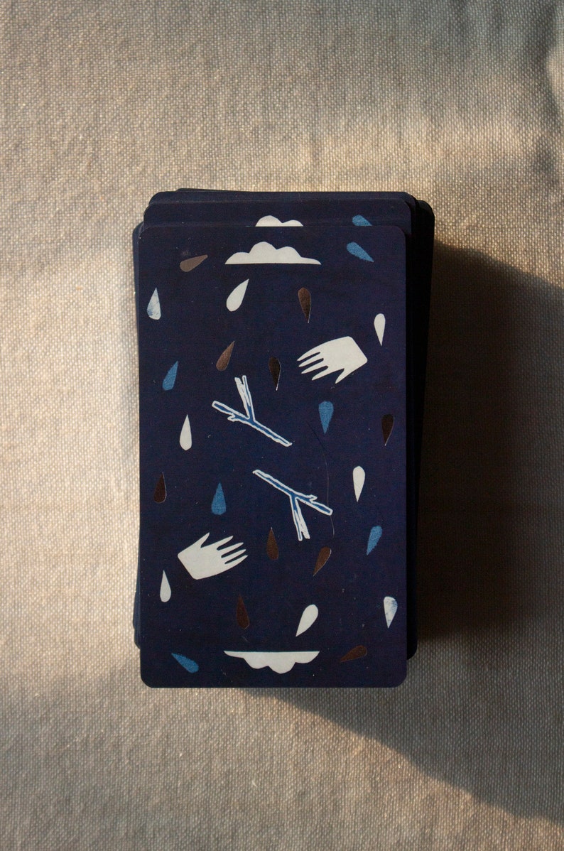 May include: A deck of cards with a navy blue background. The cards feature white hands, clouds, and raindrops in blue and brown. Two stylized branches are also present. The cards are stacked on a neutral-colored surface.