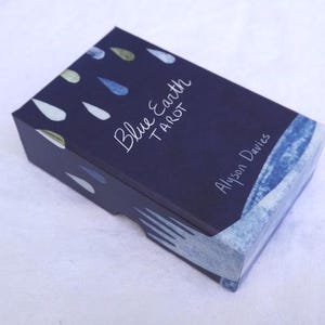 May include: A closed, rectangular tarot card box with a dark blue background and white text that reads "Blue Earth Tarot" and "Alyson Davies." The box has a watercolor design of blue and green teardrop shapes.