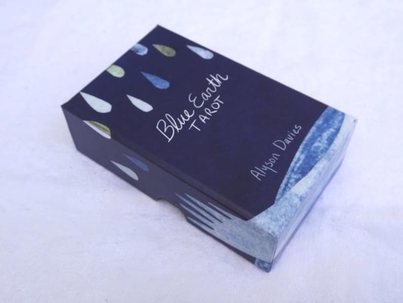 May include: A closed, rectangular tarot card box with a dark blue background and white text that reads "Blue Earth Tarot" and "Alyson Davies." The box has a watercolor design of blue and green teardrop shapes.