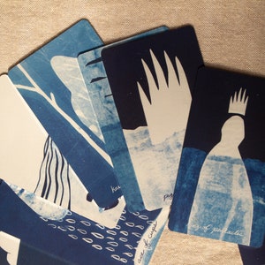 May include: A collection of tarot cards with indigo blue and white illustrations. The cards feature various designs, including abstract figures, trees, and hand-like shapes. The cards are arranged in a fan shape, with some text visible on the cards.