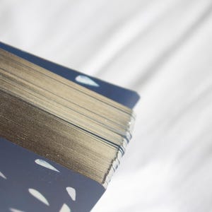 May include: A deck of cards with gilded edges, featuring a navy blue design with white illustrations of hands and teardrop shapes. The cards are stacked, showing the gold edges and the patterned back.