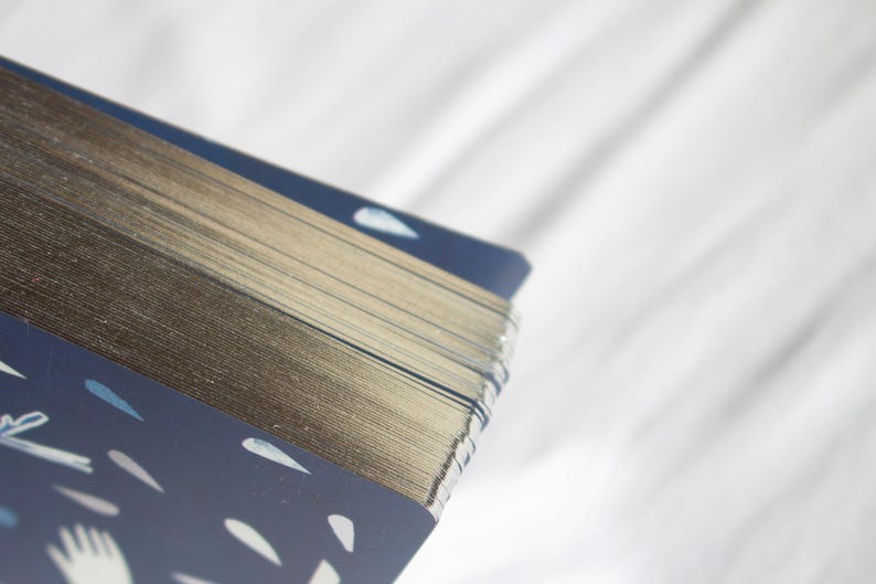 May include: A deck of cards with gilded edges, featuring a navy blue design with white illustrations of hands and teardrop shapes. The cards are stacked, showing the gold edges and the patterned back.
