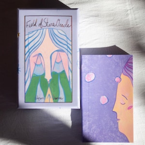Puede incluir: Un juego de cartas Oracle Field of Stars y su guía. La caja es de color lila claro con una ilustración de una cara con pelo azul y montañas verdes. La guía es morada con un perfil de una cara. El texto "Field of Stars Oracle" está en la caja.