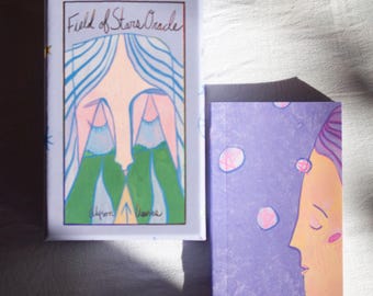 May include: A Field of Stars Oracle deck and guidebook. The box is light purple with an illustration of a face with blue hair and green mountains. The guidebook is purple with a side profile of a face. The text "Field of Stars Oracle" is on the box.