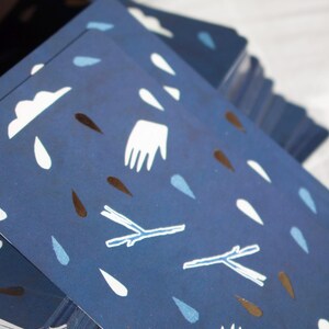 May include: A stack of navy blue cards with a repeating pattern of white clouds, raindrops, hands, and twig-like shapes in white and brown. The cards are stacked at an angle, revealing the patterned design.